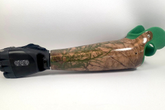 This 14ELMT custom transradial prosthesis features a laminated fabric sleeve over a carbon fiber frame featuring a woodland image and a coordinating green silicone socket. It is paired with a TASKA Hand.